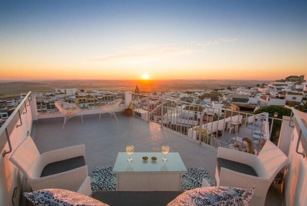 A rooftop terrace with outdoor seating, a table with glasses, and a sunset view over a cityscape.