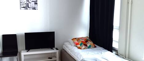 A small, neatly arranged bedroom with a single bed, a chair, a TV, and a nightstand.