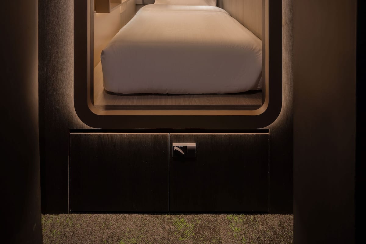 Una habitación pequeña y bien iluminada con cama, una mesita de noche y un panel de control.
