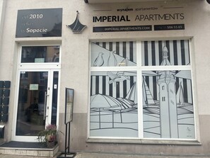 Un edificio con un letrero que dice 'Imperial Apartments' y una dirección web. Tiene un gran ventanal con un diseño a rayas blanco y negro y una ilustración de un pájaro.