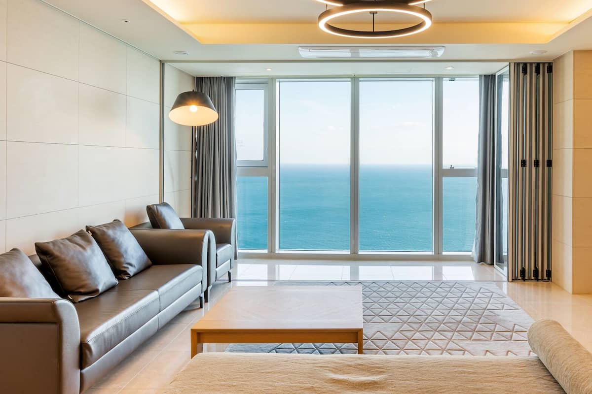 A modern living room with a sofa, coffee table, and TV, large windows with a view of the sea, and a ceiling light fixture.