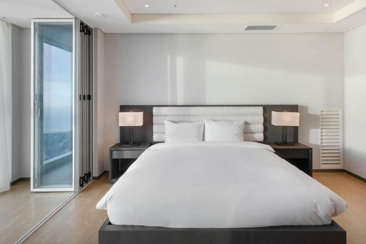A modern hotel room with a large bed, two bedside tables with lamps, a sliding glass door, and a view of the city.