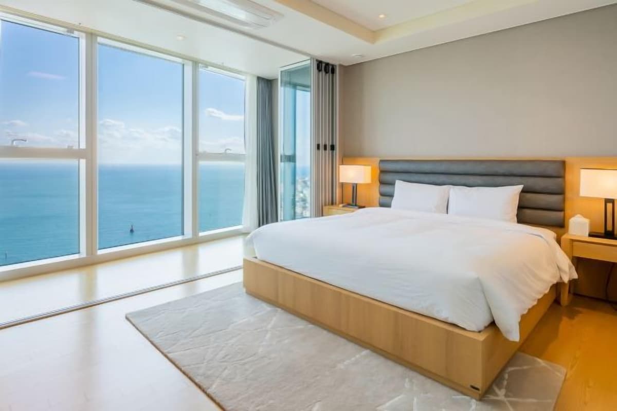 A modern bedroom with a large bed, two bedside lamps, and a view of the sea through floor-to-ceiling windows.