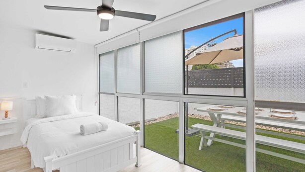 A modern bedroom with a large window, a ceiling fan, and a view of an outdoor patio. - Experience the ultimate coastal getaway in this contemporary three-bedroom unit! (Gold Coast)