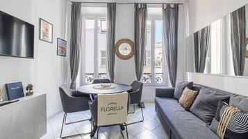 A modern living room with a grey sofa, a dining table with chairs, and a TV mounted on the wall.