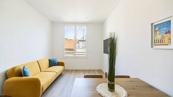 A modern living room with a yellow sofa, a wooden dining table, and a large window with a view of buildings.