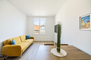 A modern living room with a yellow sofa, a wooden dining table, and a large window with a view of buildings.
