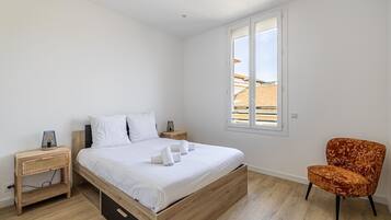 A bedroom with a wooden bed, white bedding, a wicker nightstand, a modern orange chair, and a window with a view of buildings.