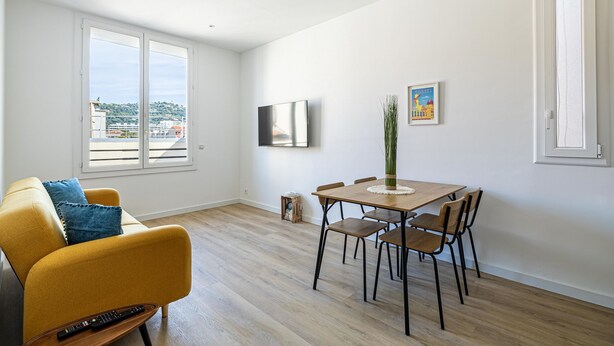 Una sala de estar moderna con un sofá amarillo, una mesa de comedor con sillas, un televisor de pantalla plana y vistas a la ciudad a través de amplios ventanales.