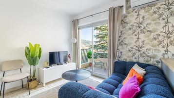 A modern living room with a blue sofa, a small round table, a TV on a white cabinet, and a balcony with a view of greenery.