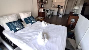 A hotel room with a bed, pillows, a bedside table, a lamp, and a view of the kitchen area.