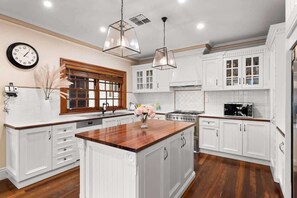 Une cuisine avec des meubles blancs, un îlot central en bois, des luminaires suspendus, une horloge et une fenêtre avec des stores.