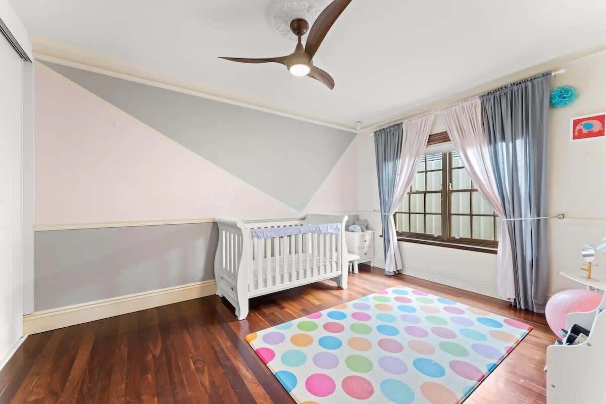 Une pièce avec un berceau blanc, un ventilateur de plafond, une fenêtre avec des rideaux et un tapis coloré.