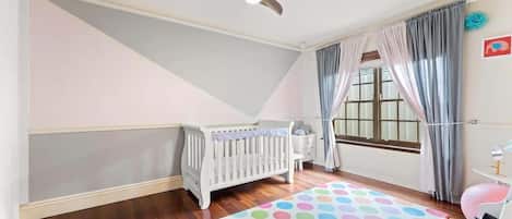 A room with a white crib, a ceiling fan, a window with curtains, and a colorful rug.