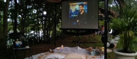 An outdoor setting with a projection screen displaying a person. There is a blanket and pillows on the ground, and a potted plant to the right.