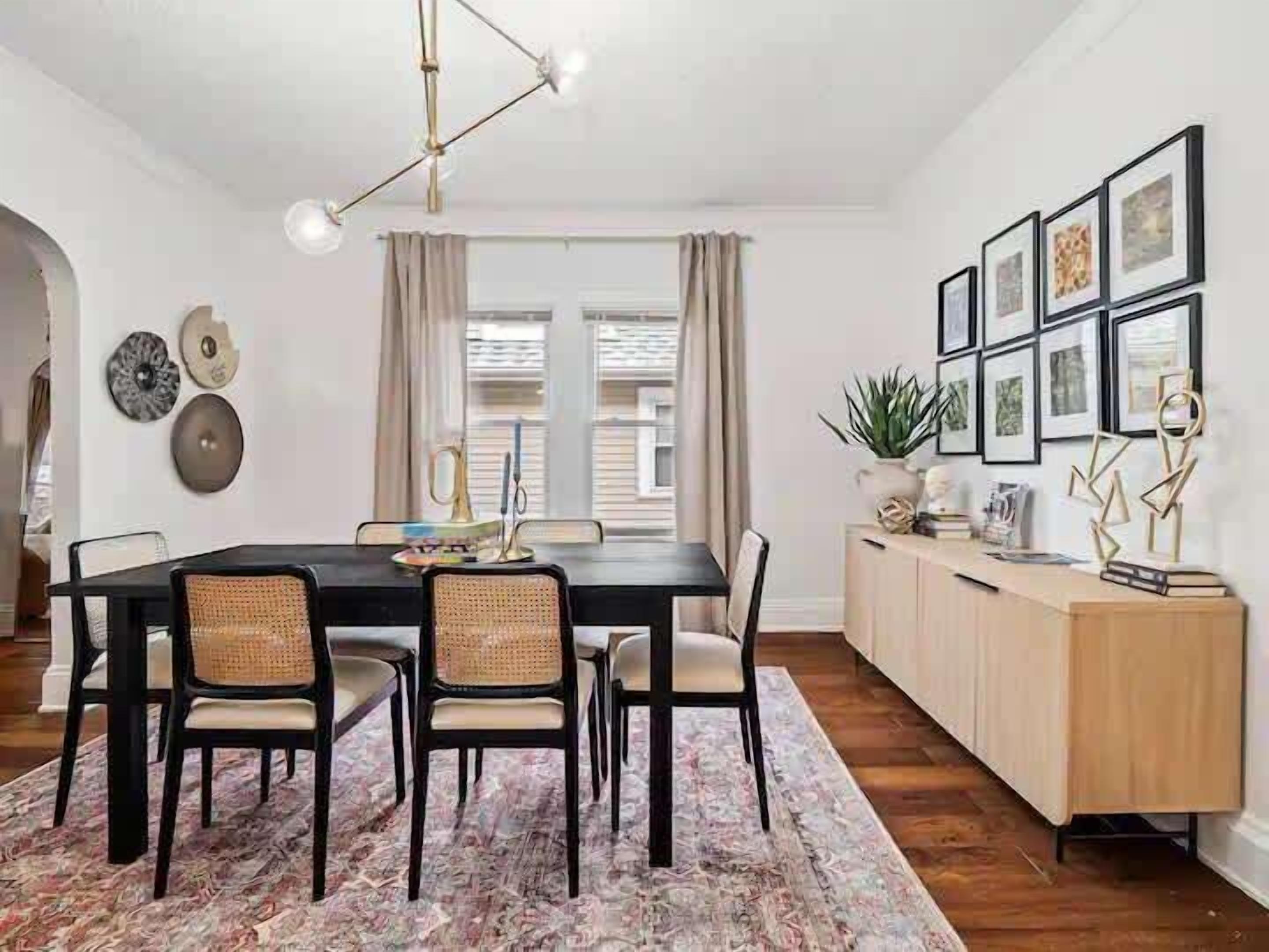 Comedor con mesa negra, cuatro sillas, un tapete estampado y un mueble de madera con marcos de cuadros.