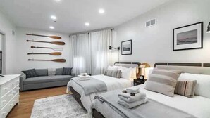 A modern hotel room with two beds, a grey sofa, a white dresser, and a wall-mounted shelf with paddles.