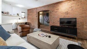 A modern living room with a brick accent wall, a flat-screen TV, a leather armchair, a wooden coffee table, and a bookshelf.