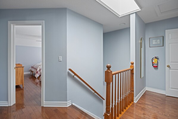 Un couloir doté d’un plancher en bois, un escalier avec une rampe en bois et une porte sur laquelle est fixé un extincteur au mur.