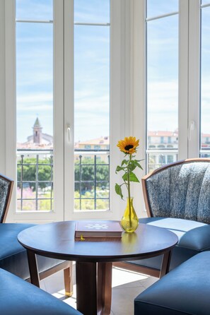 A room with a round table, a vase with a single flower, and a view of buildings through large windows.