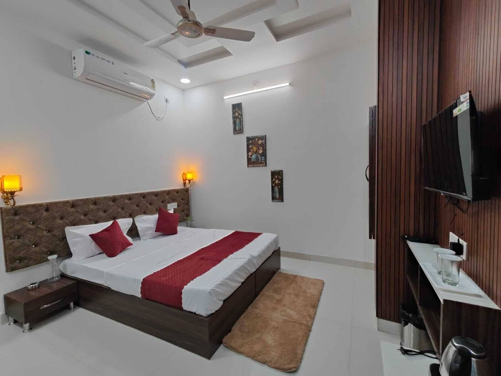 A bedroom with a bed, bedside lamps, a ceiling fan, an air conditioner, a TV, and a small desk with a chair.