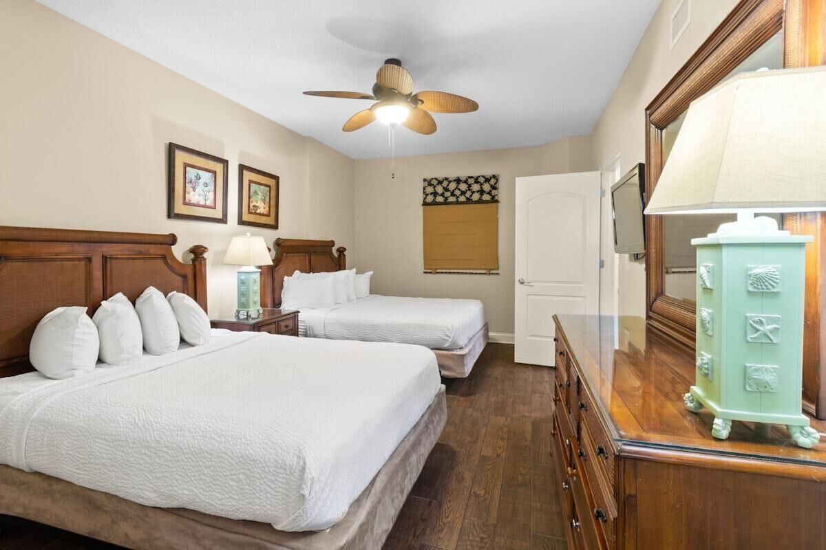 A hotel room with two beds, a ceiling fan, a wooden dresser, a mirror, and framed artwork on the wall.