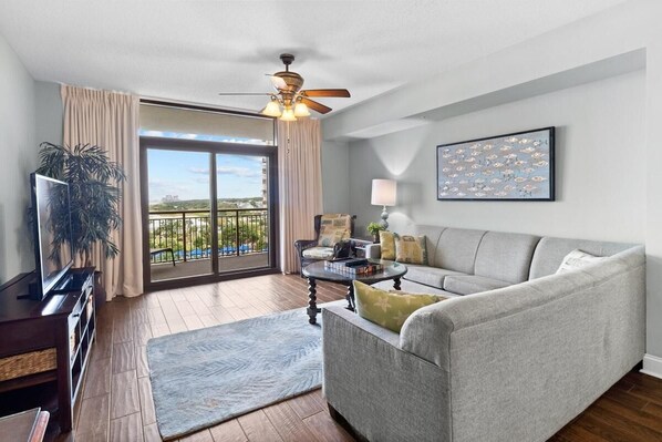 A living room with a grey sofa, a coffee table, a TV, and a ceiling fan. There is a balcony with a view of the city.