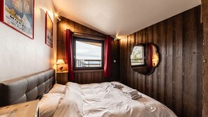 A bedroom with a wooden wall, a large window with curtains, a bed, a nightstand with a lamp, and a mirror. - Le Belvédère - IMMODREAMS - Avoriaz (Avoriaz)