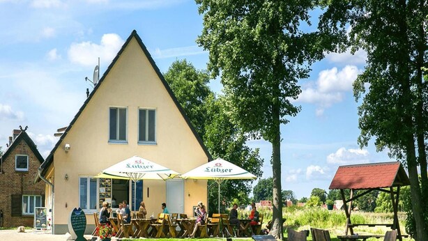 Un restaurant situé au bord de la rivière, avec une terrasse et des parasols, un canal au premier plan et un bâtiment avec un toit en pente en arrière-plan.