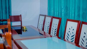 Uma sala de conferência com mesa de vidro, cadeiras de madeira com encosto em padrão de folhagem e cortinas verde-água.