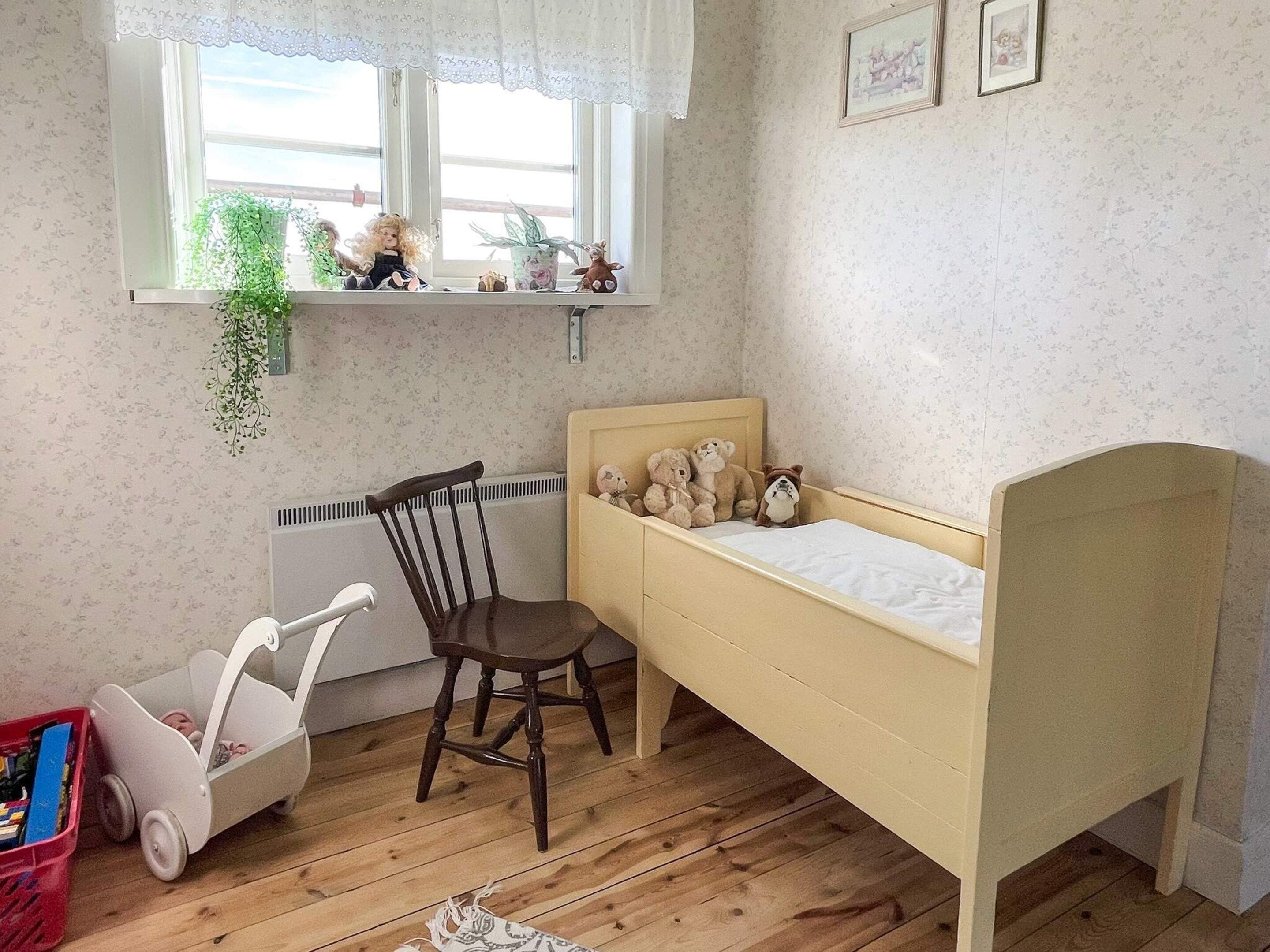 A child's bedroom with a wooden bed, a chair, a radiator, a window with plants, and a small shelf with toys.