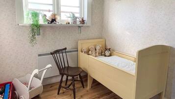 A child's bedroom with a wooden bed, a chair, a radiator, a window with plants, and a small shelf with toys.