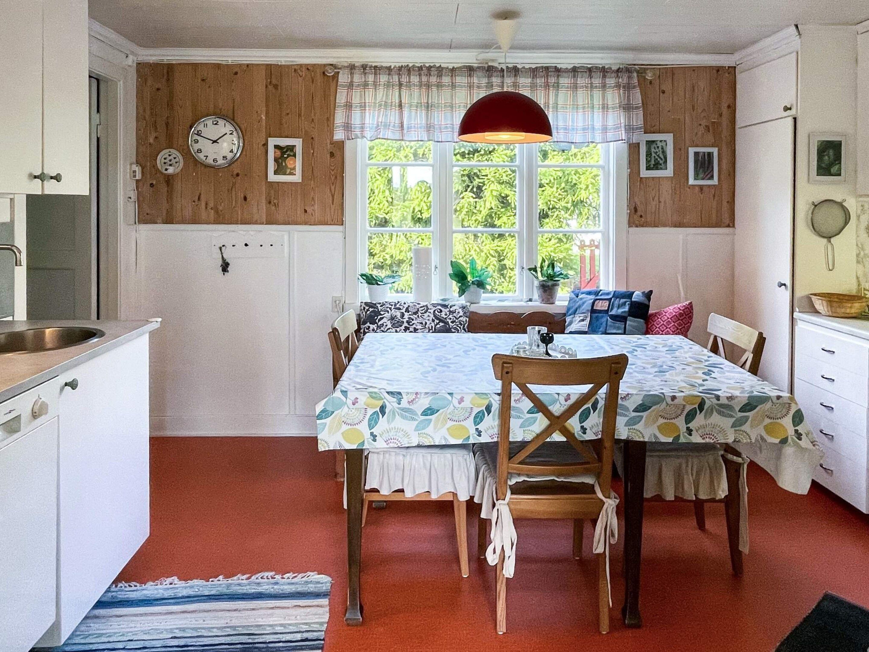 A cozy kitchen with wooden walls, a dining table with chairs, a red pendant light, and a window with plants.