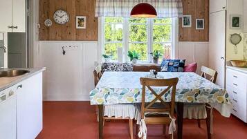 A cozy kitchen with wooden walls, a dining table with chairs, a red pendant light, and a window with plants.