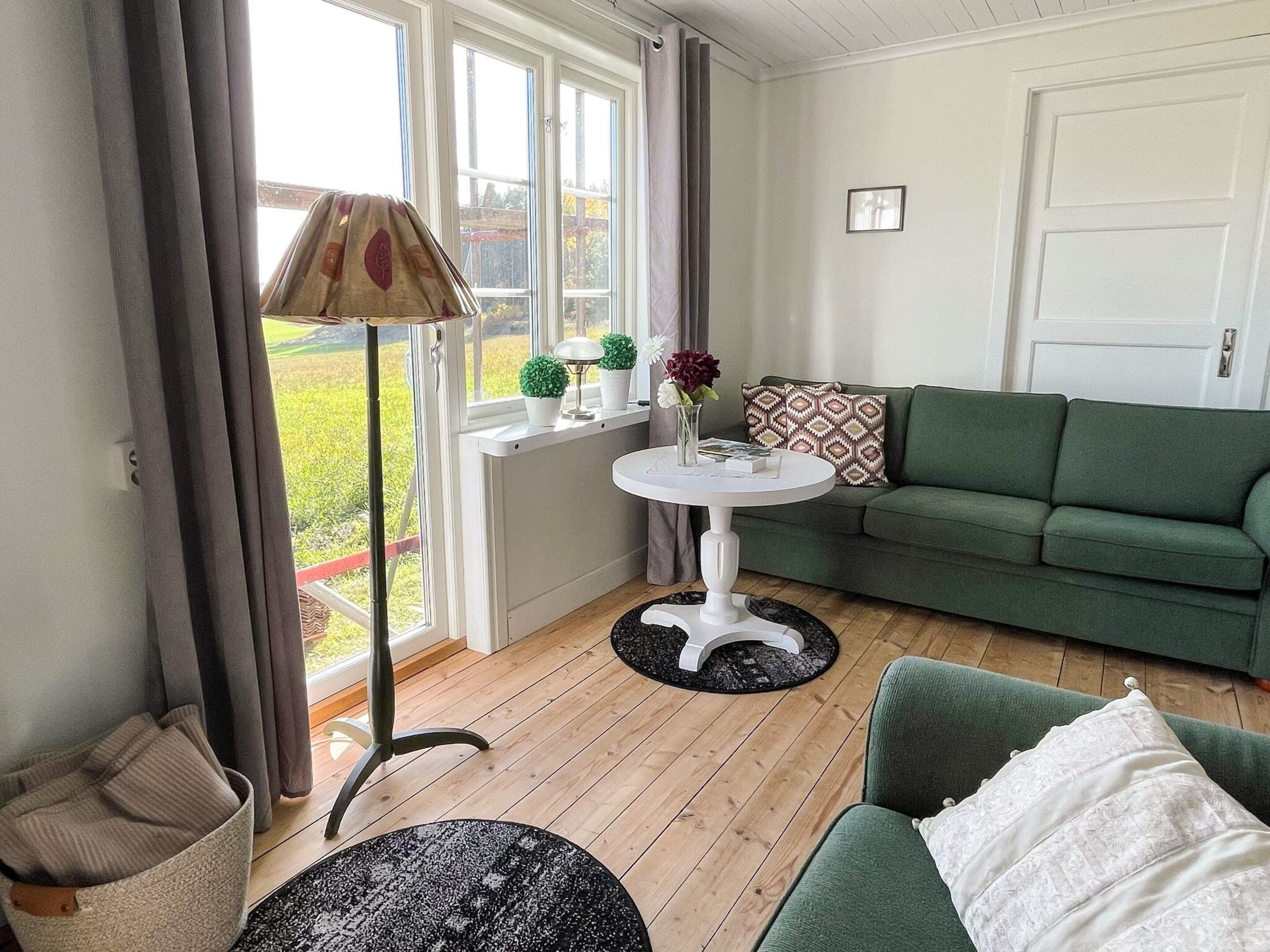 Une pièce avec un canapé vert, une petite table blanche ornée d’un vase, une lampe et une vue sur un jardin, visible à travers de grandes fenêtres.