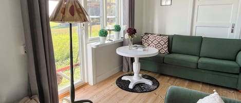 Une pièce avec un canapé vert, une petite table blanche ornée d’un vase, une lampe et une vue sur un jardin, visible à travers de grandes fenêtres.