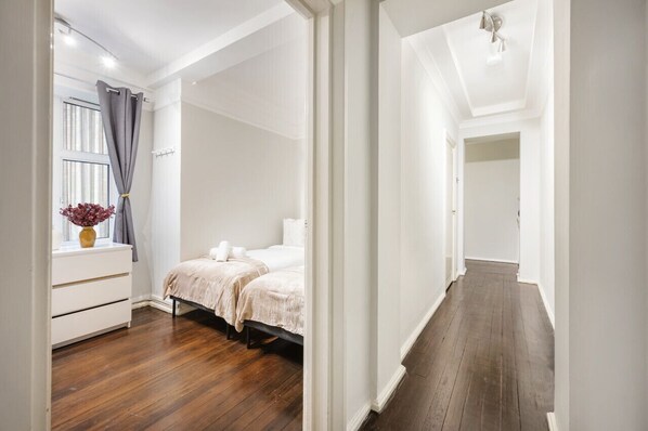 Un dormitorio con piso de madera, una cama con ropa blanca, una mesita de noche y una ventana con cortinas.
