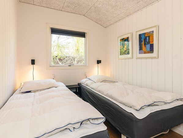 A room with two beds, a window with blinds, two wall-mounted lamps, and two framed artworks.