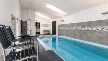 A swimming pool with black lounge chairs, a tiled floor, and a wall with a mosaic pattern.