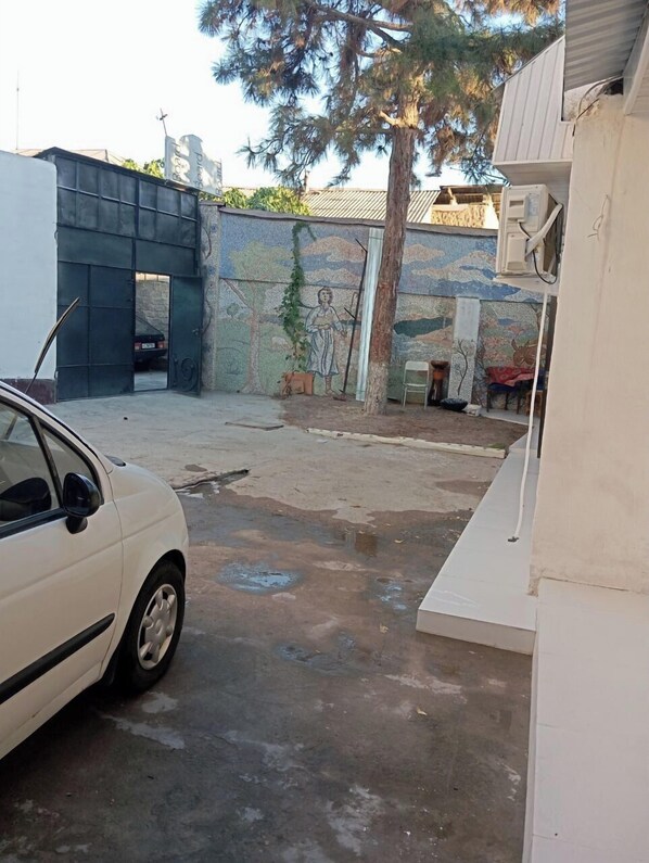 A white car parked in a driveway with a mural on the wall and a tree in the background. - Hostel Karadeniz ok (Tashkent)