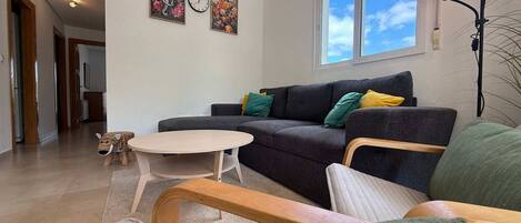 A living room with a sofa, a round table, and a small stool. There are two wall clocks and a window with a view of the sky.
