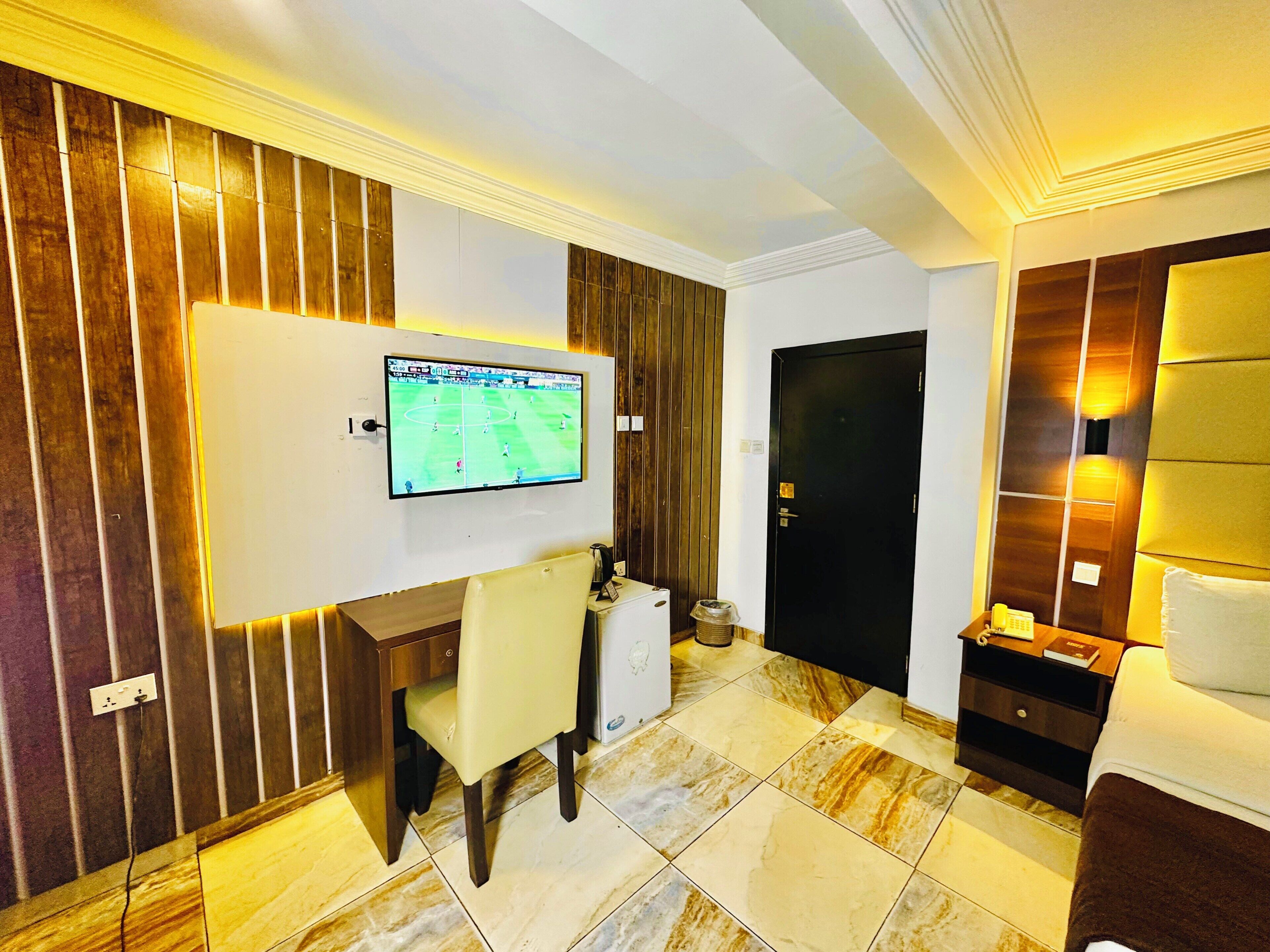 Habitación de hotel con televisión de pantalla plana montada en la pared, un escritorio con silla, una puerta negra y una mesita de noche con lámpara.