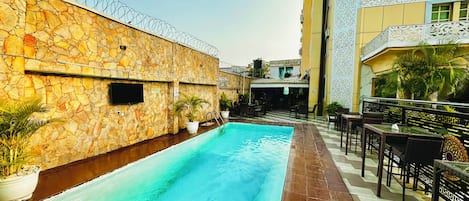 A swimming pool with a stone wall and barbed wire on one side, and a covered seating area with tables and chairs on the other.