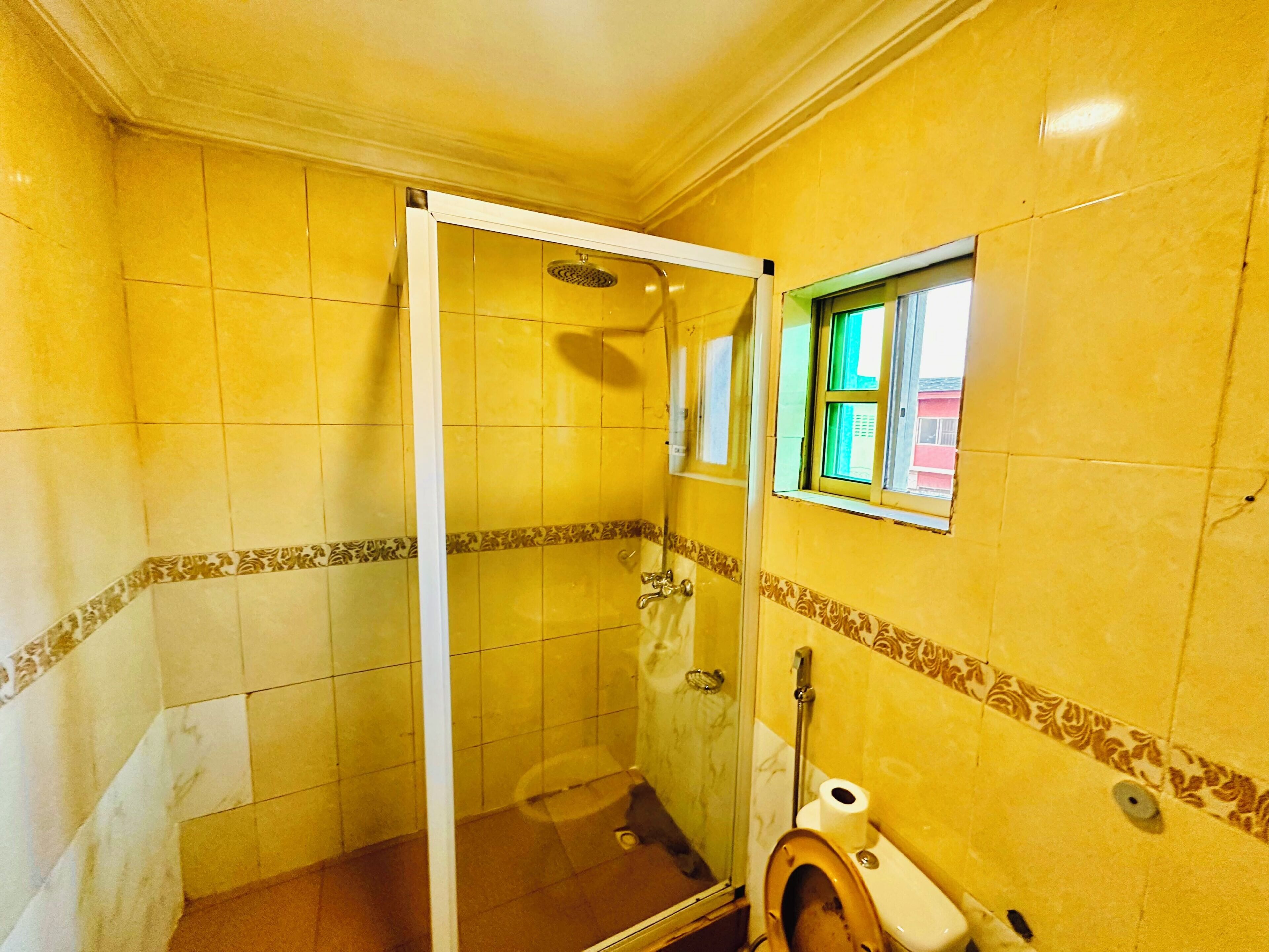 A bathroom with yellow tiled walls, a glass shower enclosure, a toilet, and a small window.