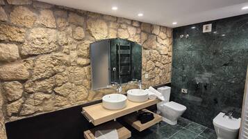 A modern bathroom with a stone wall, a wooden vanity, two sinks, a toilet, and a shower area.
