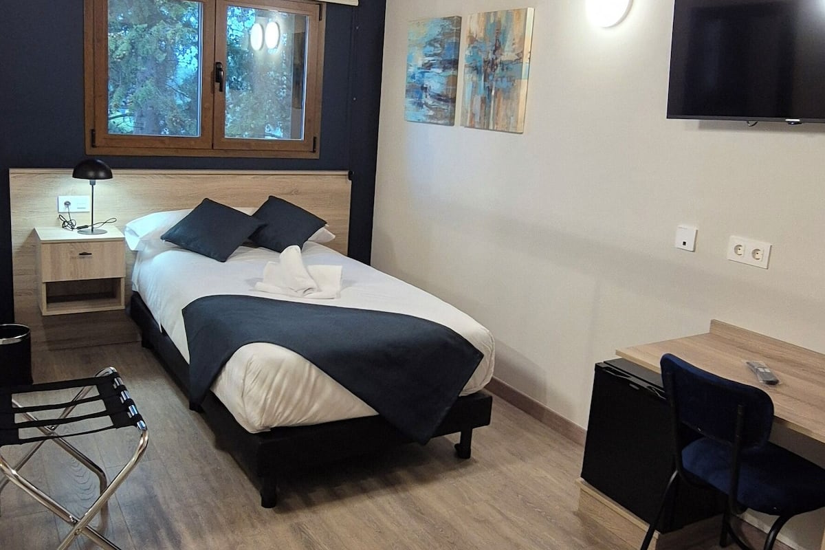 A modern bedroom with a wooden ceiling, a bed with a dark headboard, a desk with a chair, and a TV mounted on the wall.