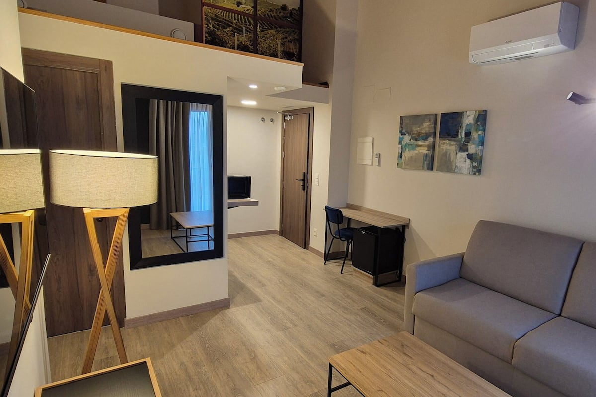 A modern living room with a sofa, a coffee table, and a TV. There is a staircase leading to an upper level, and a door to the right.