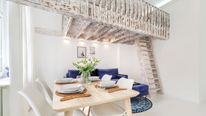 Ein Essbereich mit einem Holztisch, gedeckt für vier Personen, weißen Stühlen, einer Vase mit weißen Blumen und einem blauen Sofa. Eine Holzleiter ist an der Wand zu sehen.