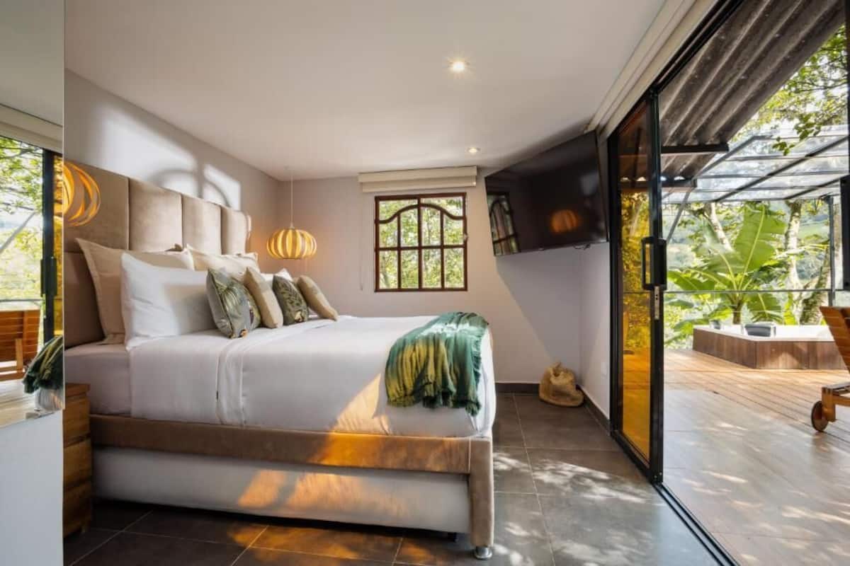 A modern bedroom with a large bed, a TV mounted on the wall, and a sliding glass door leading to an outdoor area.