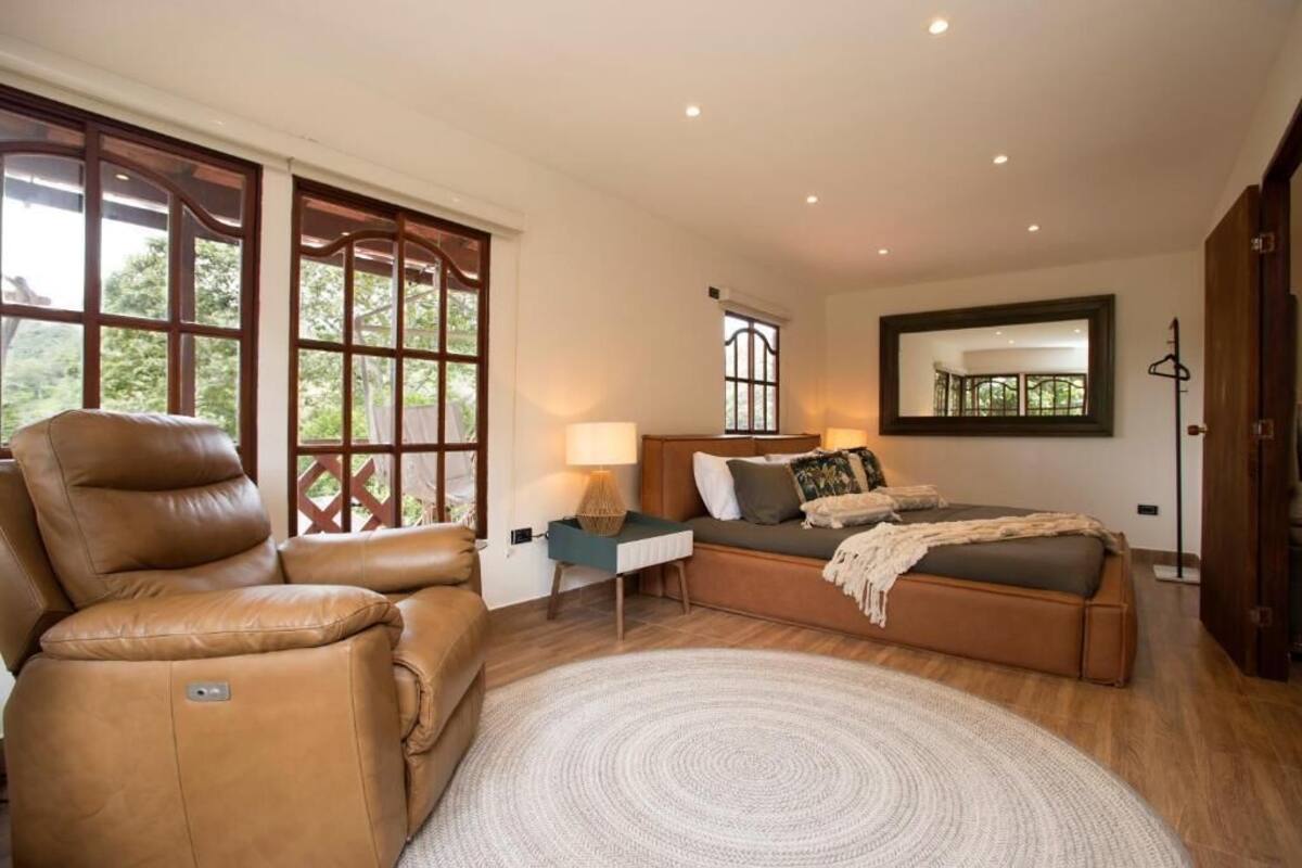 A bedroom with a sofa, a bed, a standing lamp, and a large window.